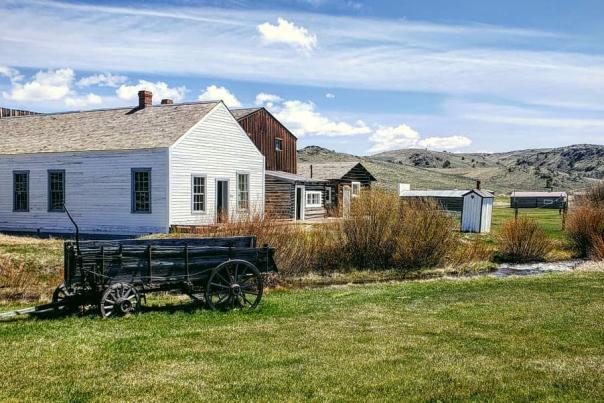 Historic wooden buildings and a vintage wagon at a frontier-style site in Wyoming, surrounded by sagebrush and open grasslands under a partly cloudy sky.