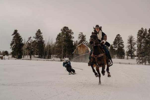 A horseback rider pulls a skier through the snow during a skijoring run near Wyoming. The horse gallops across a snow-covered field as the skier follows behind, with a rustic wooden barn, fencing, and evergreen forest creating a classic Wyoming winter setting.