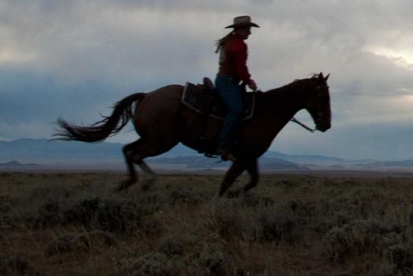 A silhouetted horseback rider in a hat rides into the blazing sunset beneath a cloudy sky, mountains in the far distance.