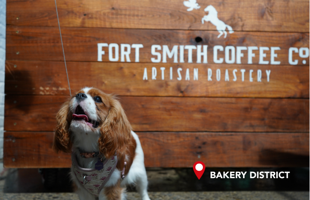 A happy dog looks at her owner in front of the Fort Smith Coffee Co. sign.