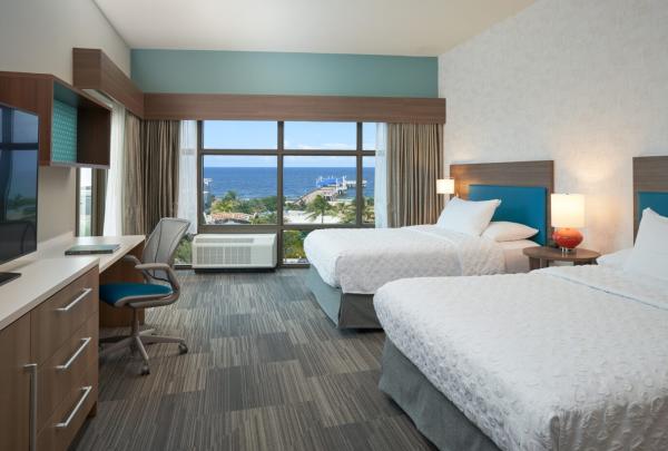 An hotel room with 2 queen beds, a desk, and a console table with drawers, all with an ocean view of Pompano Beach and the Fisher Family Pier