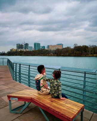 Two young children sitting on a bench on the Hike & Bike Trail Boardwalk. Lady Bird Lake and the Austin skyline are behind them