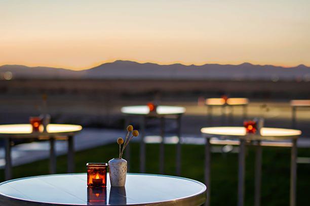 6 small metal cocktail-sized tables spread out on a lawn, each with a small square red candle and a small white vase with florals centered on top. Mountains are blurred in the background with a sunset sky.