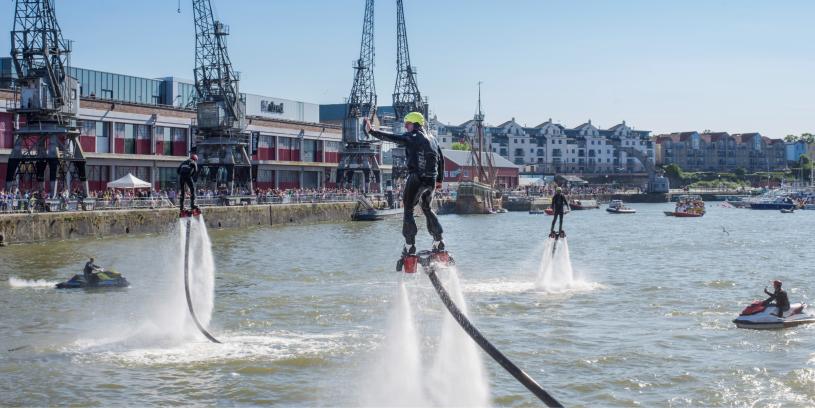 A man wearing a water-powered jetpack performing stunts on Bristol's Harbourside during the Bristol Harbour Festival with the M Shed local history museum in the background - credit Jim Cossey
