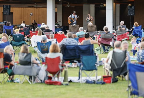 Guests relax in lawn chairs on the grass during a Ditto Landing Summer Sunset Concert Series performance. A duo performs on stage while families and friends enjoy live music along the Tennessee River in a laid-back outdoor setting.