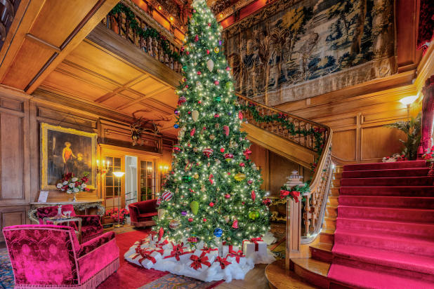 Lavishly decorated room with a large Christmas tree, red carpeted stairs, ornate wooden paneling, and elegant furniture.