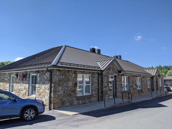 A gray stone building, the Watauga County Courthouse Annex Building, with a row of windows and a door facing the parking lot. A blue car is visible on the left side of the photo.