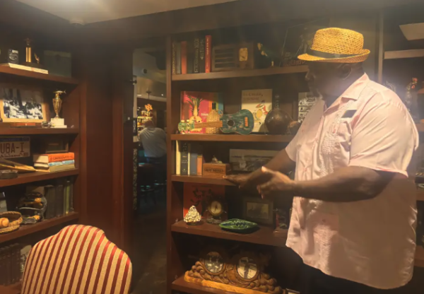 A large man in a Panama hat points to an entry way behind a sliding bookcase