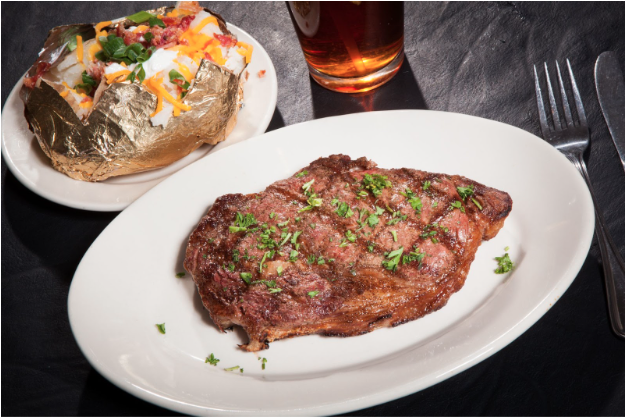 A steak grilled and charred with parsley plated with a baked potato, drink, and fork and knife.