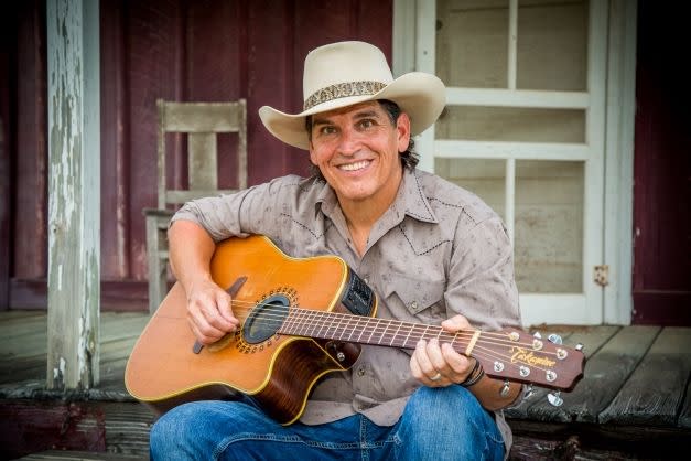 A man in a western shirt and light cowboy hat smiles at the camera as he strums an acoustic guitar from the porch of a rustic red-and-white house.