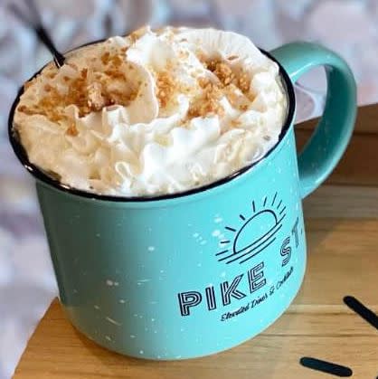 Image is of a blue, coffee mug that say's "Pike Street" on it and inside is a cinnamon roll latte with whipped cream and streusel bits.