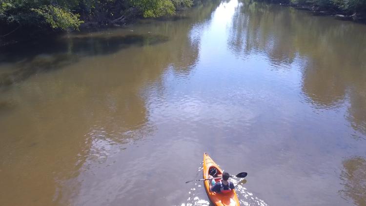 A kayaker paddles a river in the sun captured by a drone.