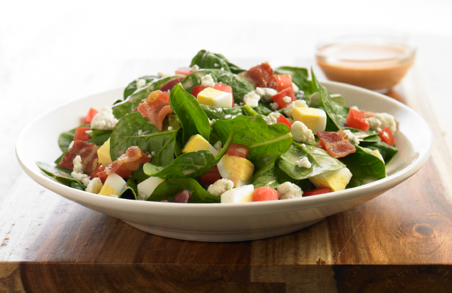 Image of a table with a white bowl filled with a green salad that has tomato's, bacon and cut up egg in it.