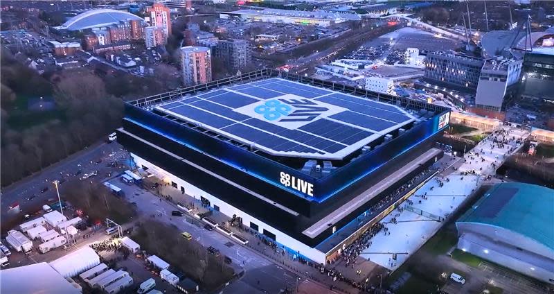 Aerial view of the Co-op Live arena in Manchester at dusk, showing solar panels on the roof and people gathering outside the venue.