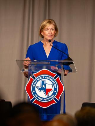 Visit The Woodlands Chairman of the Board Linda Nelson speaks at the Texas State Association of Fire Fighters 49th Biennial Convention. Nelson is in a bright blue dress, standing behind a podium bearing the TSAFF seal/emblem/logo.