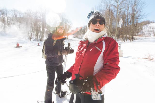 Two women are bundled in snow gear and wearing snowshoes holding hiking poles and laughing.