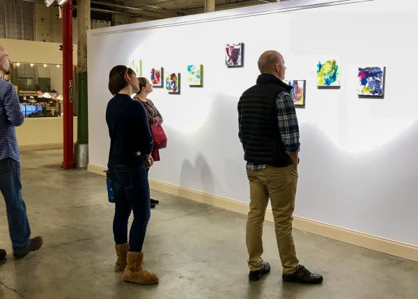 Visitors view a gallery exhibition at Lowe Mill ARTS & Entertainment in Huntsville. Several people stand in an industrial-style gallery space, observing colorful abstract paintings displayed on a large white wall.