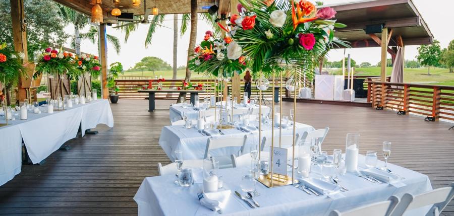 Seven rectangular tables of 6 set with white linen table cloths and red napkins against the sun setting over the Pompano Beach Golf Course