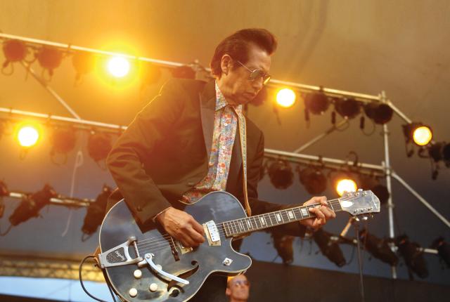 Alejandro Escovedo with shades and a guitar, live and in concert with lights on stage.