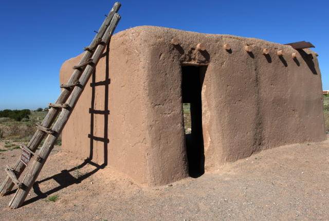 Small adobe building with a wooden ladder leaning against its side under a clear blue sky.