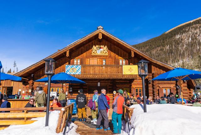 A rustic wooden lodge in a snowy mountain setting, with people gathered outside under blue umbrellas, enjoying the sunny weather.