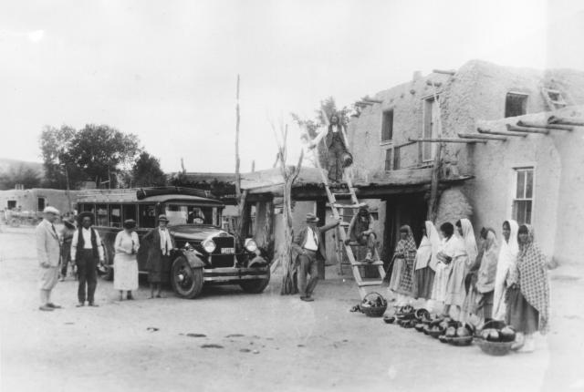 Historic Pueblo scene with an early motor bus and people near multi‑story adobe buildings.