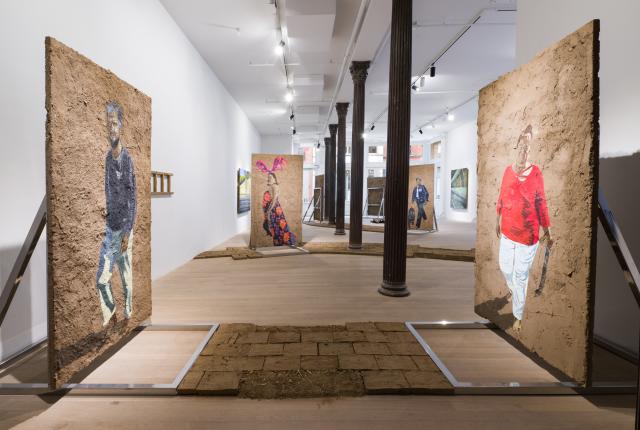A contemporary gallery installation features large textured panels resembling earthen walls, each painted with full‑body figures, arranged along a central path of brick-like tiles under bright overhead lighting.
