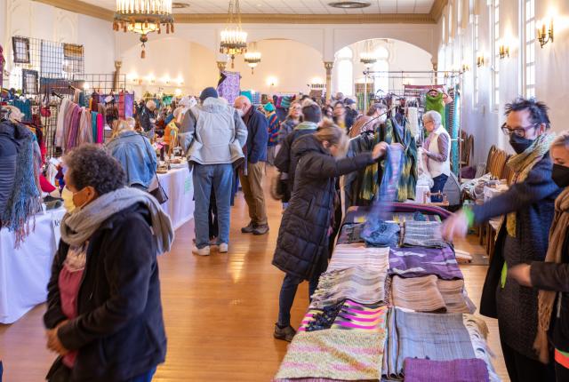 A bustling indoor craft fair, with people in winter clothing browsing colorful textile displays.