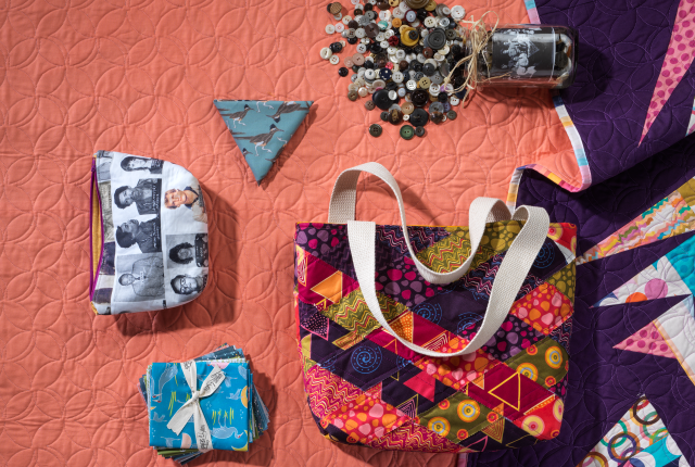 Colorful quilting display on a textured orange backdrop. Includes multicolored quilt, patterned bags, a stack of fabrics, buttons, and a jar.