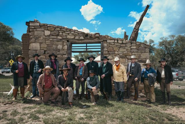 Group of people dressed in Old West–style clothing posing in front of a partially collapsed stone building under a bright blue sky.