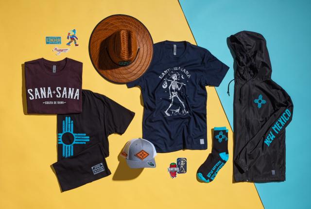 A flat lay of New Mexico–themed apparel and accessories displays graphic T‑shirts, a wide‑brimmed hat, a jacket, socks, a cap, and colorful stickers arranged on a split yellow and turquoise background.