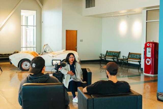 A camera crew films an interview in a spacious room that features a vintage white convertible car, several benches, and a red Texaco gas pump.
