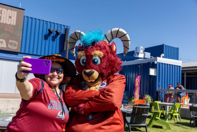 A fan holds up a purple phone for a selfie with a red goat mascot wearing a hockey jersey.