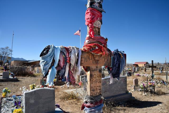 A wooden cross, draped with colorful scarves and bandanas, stands in a cemetery under a clear blue sky.