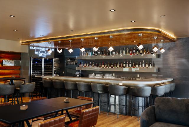 Modern bar interior with a long counter, shelves stocked with bottles, stylish lighting, and seating throughout the room.
