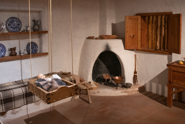 Historic-style room with a hanging cradle, adobe fireplace, wooden shelves, and rustic furnishings.