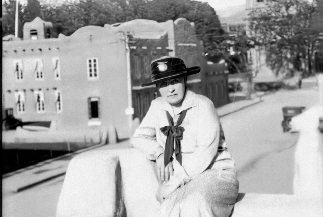 Historic black‑and‑white photo of a person sitting on an adobe wall with Pueblo‑style buildings in the background.
