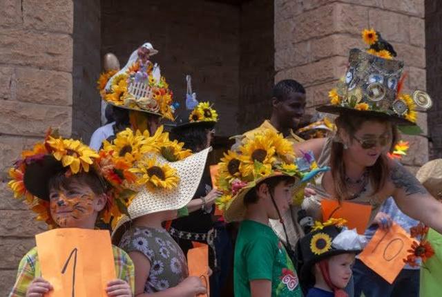 Group of people, including children, wearing decorated hats covered in sunflowers and other ornaments at an outdoor event.