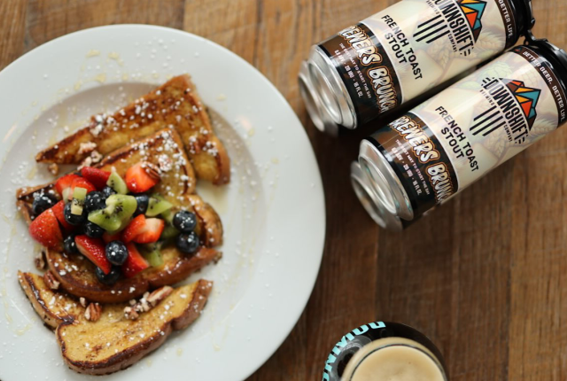 A plate of french toast topped with fruit and powdered sugar sits next to two cans of French Toast Stout and the top of a glass of stout.