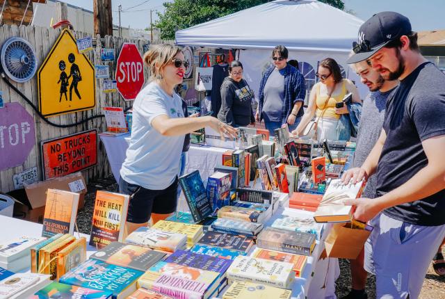 Outdoor book stall with tables full of novels and signs on a wooden fence, while people browse under a canopy.