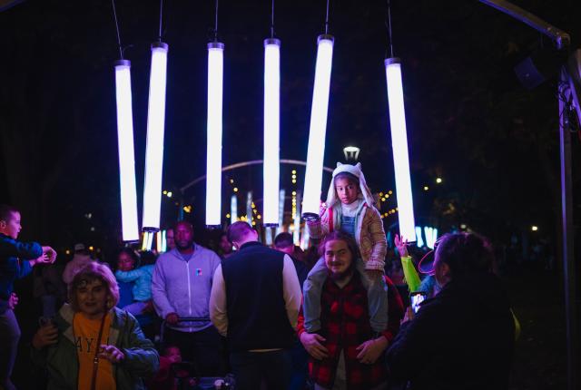 People walk beneath glowing vertical light installations at the Paseo Project in Taos, with a child on an adult's shoulders enjoying the interactive art.