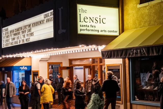 Nighttime scene outside The Lensic Performing Arts Center. A crowd of people in jackets walks beneath a marquee displaying upcoming event names.