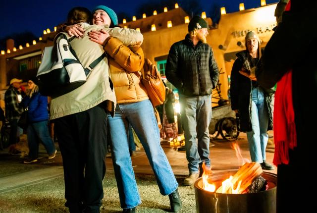 Two people warmly embrace in a festive outdoor setting with a fire pit and soft lights.