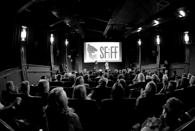 A black and white photo of an audience in a theater facing a screen that says "SFIFF" as two people stand on stage.