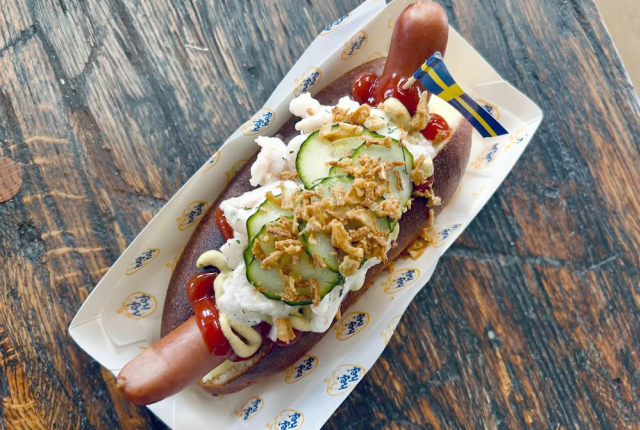 A hot dog topped with ketchup, mustard, cucumbers, chopped onions, and a Swedish flag toothpick in a tray on a rustic wooden table.