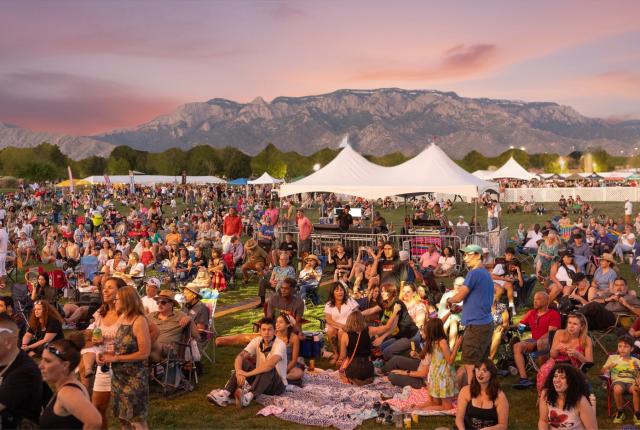 A large outdoor crowd sits on blankets and folding chairs at a festival on a grassy field, with white event tents scattered throughout and a mountain range glowing in the pink sunset sky behind them.