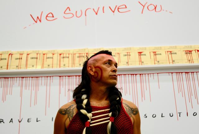 Indigenous man with braided hair and face paint stands solemnly.