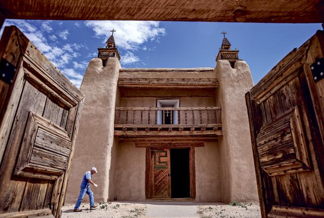 Wooden doors open toward an adobe church with twin towers and a small balcony, as a person walks across the sunlit courtyard under a bright blue sky."