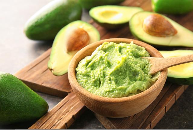 Wooden board with bowl of delicious guacamole and ripe avocados on table.