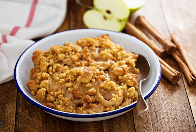 Apple crisp in a round white bowl surrounded by whole cinnamon sticks and apples.